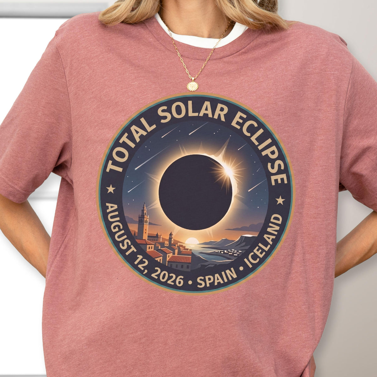 May include: A dusty rose short-sleeve t-shirt featuring a circular graphic of a total solar eclipse over a coastal town. The text reads TOTAL SOLAR ECLIPSE AUGUST 12, 2026 SPAIN ICELAND. The design includes a dark sun, a bright corona, and stars.