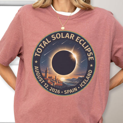 May include: A dusty rose short-sleeve t-shirt featuring a circular graphic of a total solar eclipse over a coastal town. The text reads TOTAL SOLAR ECLIPSE AUGUST 12, 2026 SPAIN ICELAND. The design includes a dark sun, a bright corona, and stars.