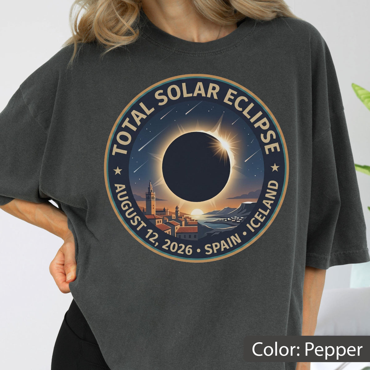 May include: A pepper-colored t-shirt featuring a circular graphic of a total solar eclipse over a coastal town. The text reads TOTAL SOLAR ECLIPSE AUGUST 12, 2026 SPAIN ICELAND. The design includes a dark sun, a bright corona, and stars.