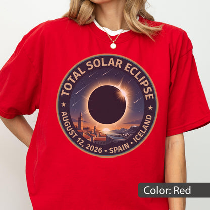May include: A red t-shirt featuring a circular graphic of a total solar eclipse over a coastal town. The text reads TOTAL SOLAR ECLIPSE AUGUST 12, 2026 SPAIN ICELAND. The design includes a dark sun silhouette, a bright corona, and stars.