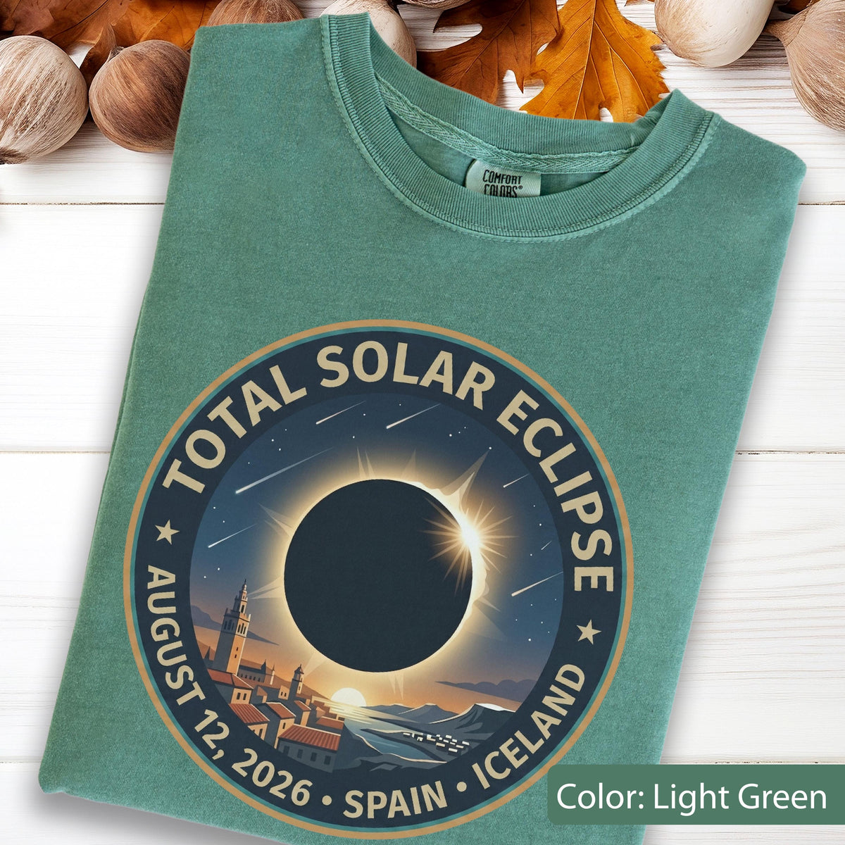 May include: A light green t-shirt featuring a circular graphic of a total solar eclipse over a town. The text reads: TOTAL SOLAR ECLIPSE, AUGUST 12, 2026, SPAIN, ICELAND. The design includes a dark moon, a bright sun corona, and falling stars.