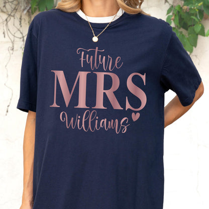 May include: A navy blue short-sleeved t-shirt featuring the text Future MRS Williams in a metallic rose gold font. The word Future and the name Williams are written in a cursive script, while MRS is in a large serif font with a small heart.