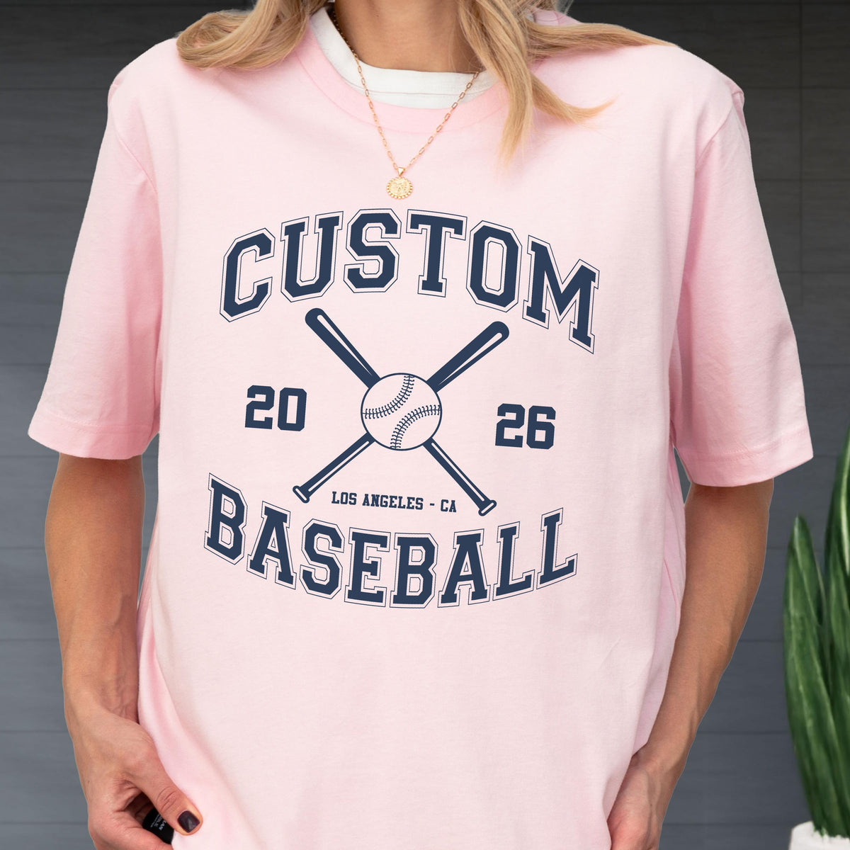 May include: A light pink short-sleeved t-shirt featuring a dark blue graphic print. The design includes the text CUSTOM BASEBALL, the numbers 20 and 26, crossed baseball bats, a baseball, and the text LOS ANGELES - CA in a collegiate font.