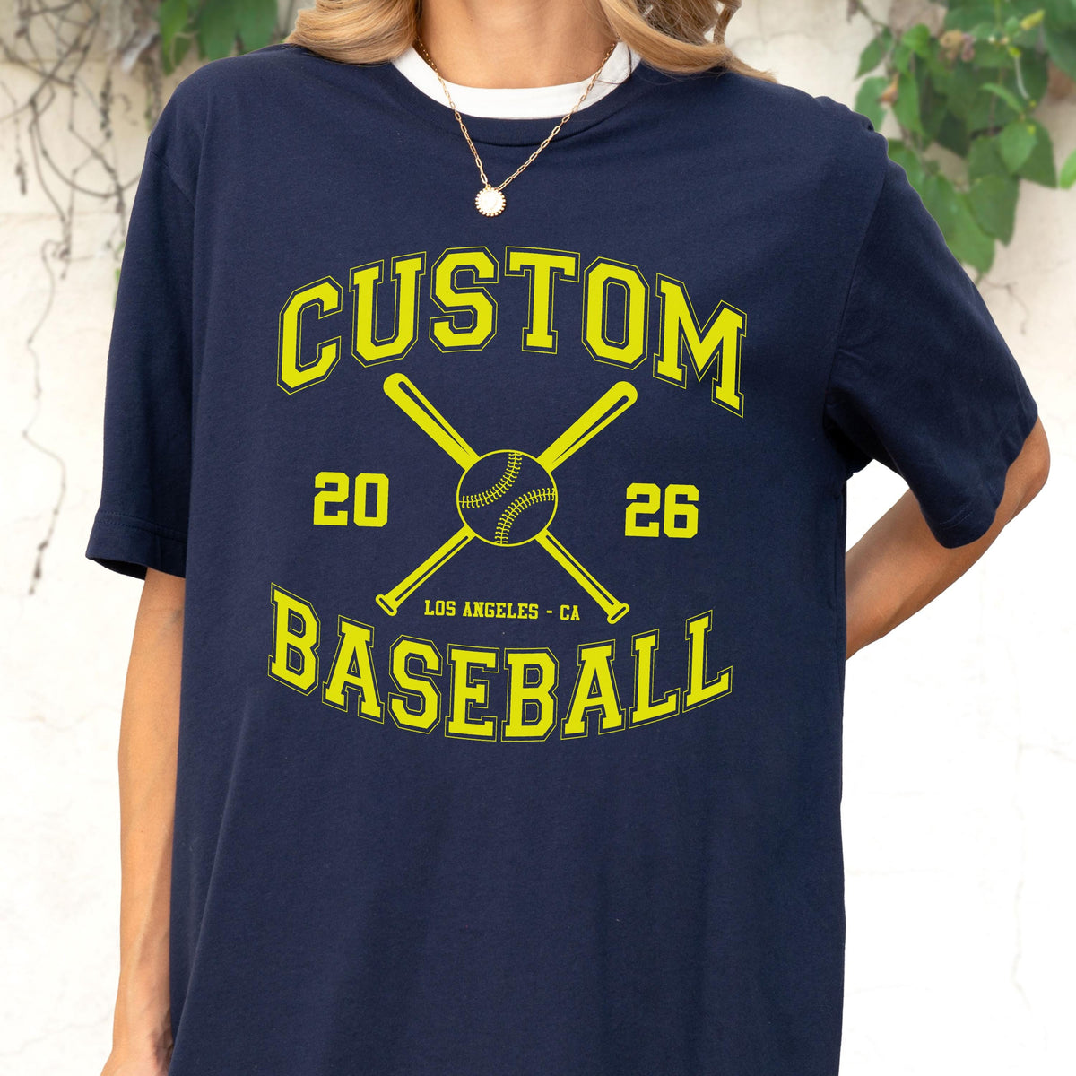 May include: A navy blue t-shirt featuring a yellow graphic print with the text CUSTOM BASEBALL, the numbers 20 and 26, and the location LOS ANGELES - CA. The design includes a central baseball icon positioned between two crossed baseball bats.