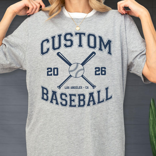 May include: A heather gray t-shirt featuring a dark blue graphic with the text CUSTOM BASEBALL, the numbers 20 and 26, and the location LOS ANGELES - CA. The design includes an illustration of a baseball centered between two crossed baseball bats.