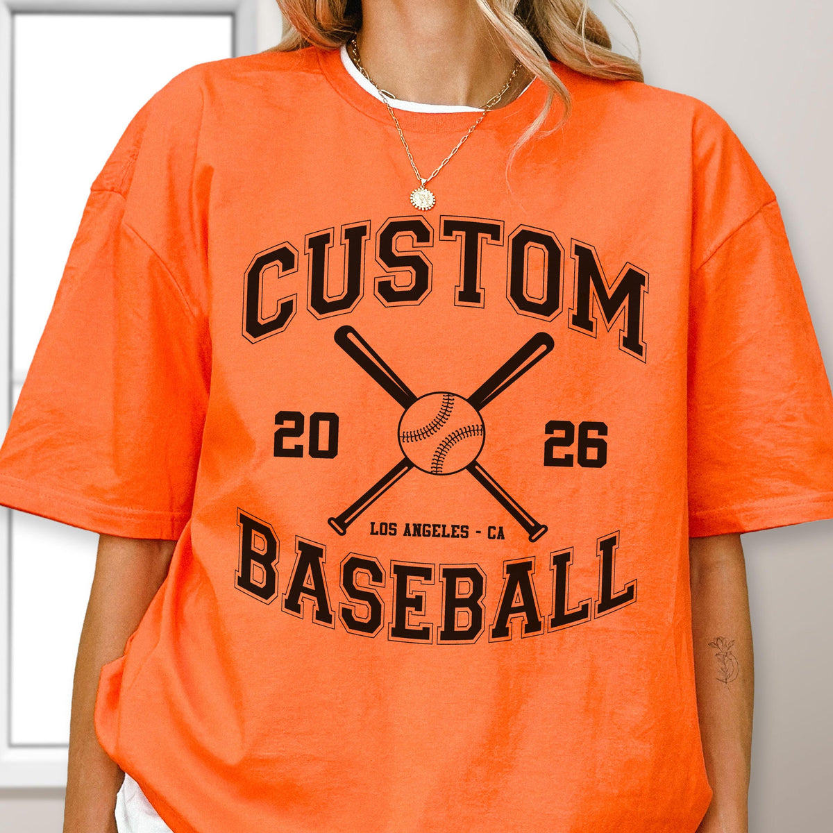 May include: A bright orange t-shirt featuring a black graphic print. The design includes the text CUSTOM BASEBALL, the numbers 20 and 26, and an illustration of two crossed baseball bats with a baseball in the center and LOS ANGELES - CA below.