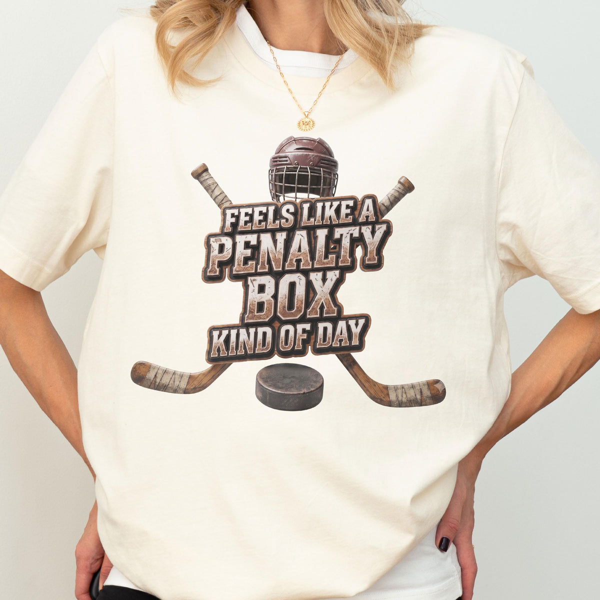 May include: A cream-colored t-shirt featuring a graphic of a brown hockey helmet, two crossed hockey sticks, and a black puck. The text reads, "FEELS LIKE A PENALTY BOX KIND OF DAY" in a distressed, block-style font centered on the chest.