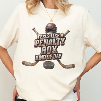 May include: A cream-colored t-shirt featuring a graphic of a brown hockey helmet, two crossed hockey sticks, and a black puck. The text reads, "FEELS LIKE A PENALTY BOX KIND OF DAY" in a distressed, block-style font centered on the chest.