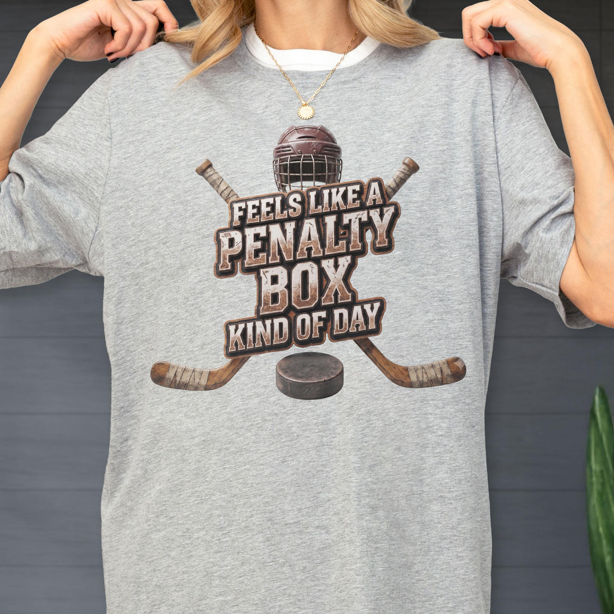 May include: A heather gray t-shirt featuring a graphic of a brown hockey helmet, two crossed hockey sticks, and a black puck. The text reads FEELS LIKE A PENALTY BOX KIND OF DAY in a distressed brown font centered over the hockey equipment.