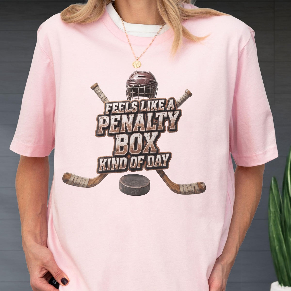 May include: A light pink short-sleeve t-shirt featuring a graphic of a hockey helmet, two crossed hockey sticks, and a puck. The text reads FEELS LIKE A PENALTY BOX KIND OF DAY in a distressed brown font centered on the chest of the garment.