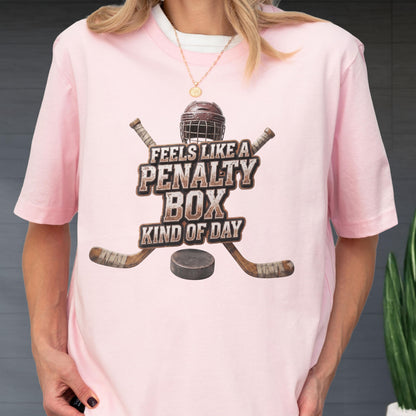May include: A light pink short-sleeve t-shirt featuring a graphic of a hockey helmet, two crossed hockey sticks, and a puck. The text reads FEELS LIKE A PENALTY BOX KIND OF DAY in a distressed brown font centered on the chest of the garment.