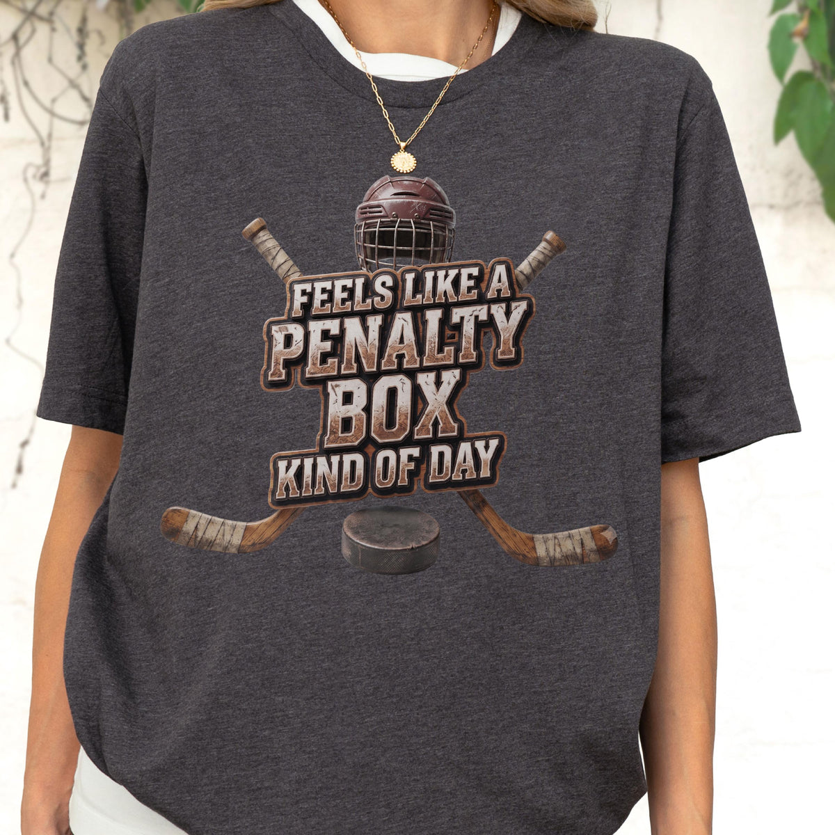 May include: A dark heather gray t-shirt featuring a graphic of a hockey helmet, two crossed hockey sticks, and a puck. The text reads FEELS LIKE A PENALTY BOX KIND OF DAY in a distressed, block-style font centered on the chest of the garment.