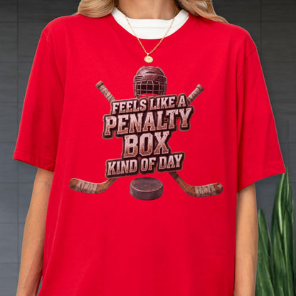 May include: A bright red t-shirt featuring a graphic of a hockey helmet, two crossed hockey sticks, and a puck. The text reads FEELS LIKE A PENALTY BOX KIND OF DAY in a distressed brown font. The shirt has a crew neckline and short sleeves.