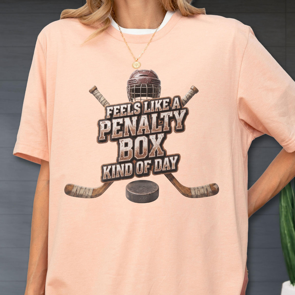 May include: A peach-colored short-sleeve t-shirt featuring a graphic of a hockey helmet, two crossed hockey sticks, and a puck. The text reads, "FEELS LIKE A PENALTY BOX KIND OF DAY" in a distressed, brown, block-style font centered on the chest.