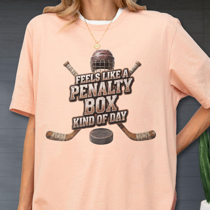 May include: A peach-colored short-sleeve t-shirt featuring a graphic of a hockey helmet, two crossed hockey sticks, and a puck. The text reads, "FEELS LIKE A PENALTY BOX KIND OF DAY" in a distressed, brown, block-style font centered on the chest.