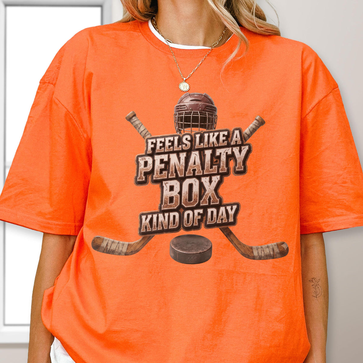 May include: An orange short-sleeve t-shirt featuring a graphic of a hockey helmet, two crossed hockey sticks, and a puck. The text reads FEELS LIKE A PENALTY BOX KIND OF DAY in a distressed brown font centered on the chest of the garment.