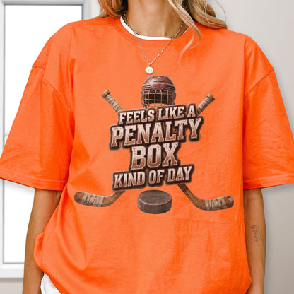 May include: An orange short-sleeve t-shirt featuring a graphic of a hockey helmet, two crossed hockey sticks, and a puck. The text reads FEELS LIKE A PENALTY BOX KIND OF DAY in a distressed brown font centered on the chest of the garment.