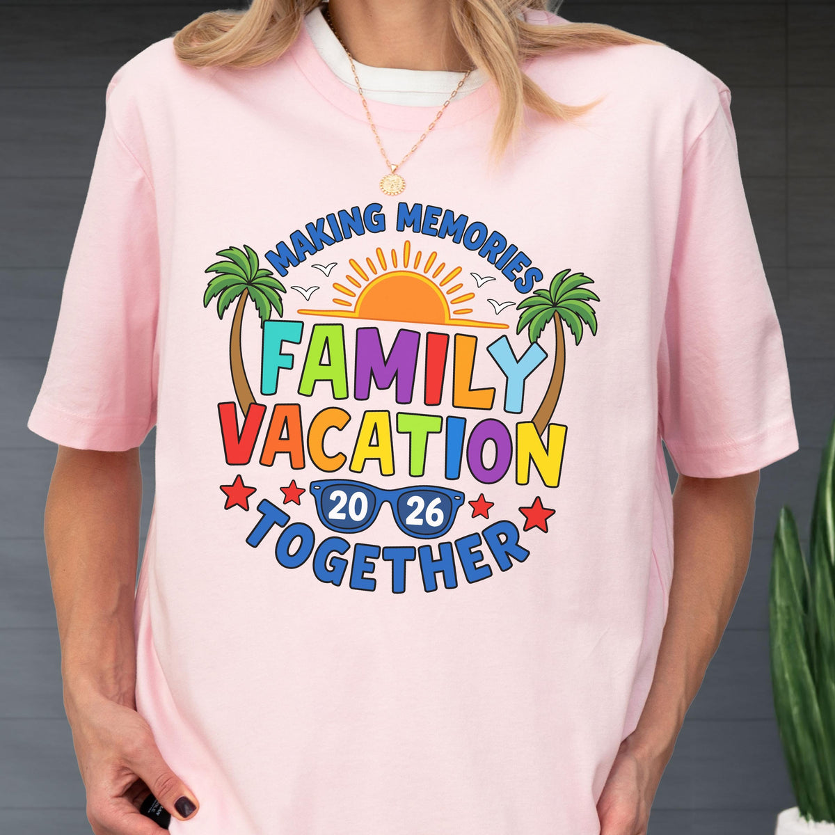 May include: A light pink t-shirt featuring a colorful graphic with the text "MAKING MEMORIES FAMILY VACATION TOGETHER 20 26". The design includes a bright orange sun, two green palm trees, blue sunglasses, and small red stars on a light pink background.