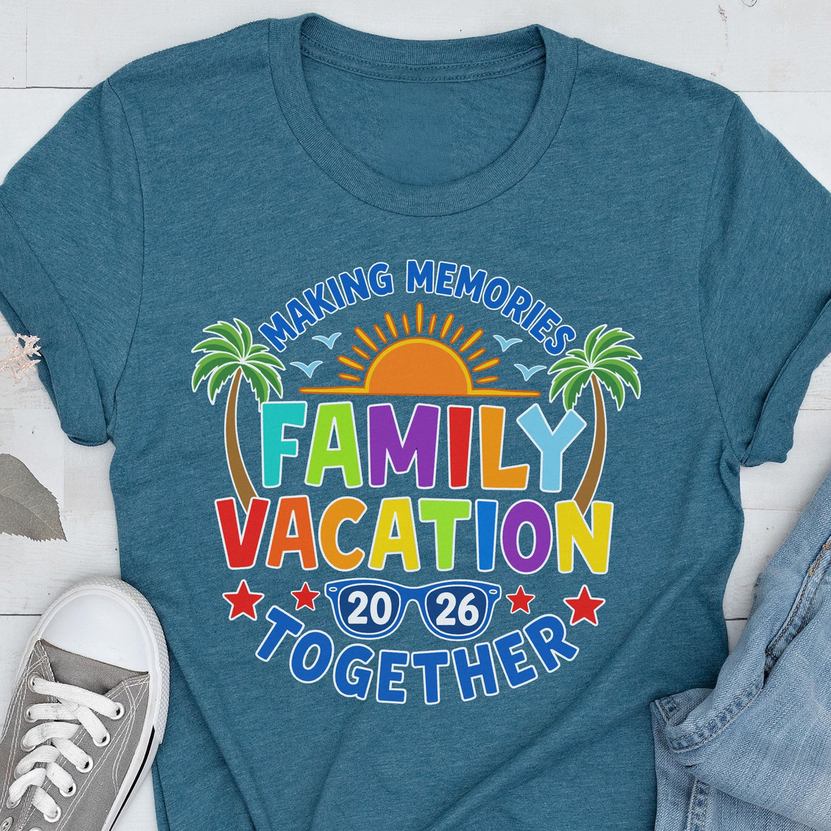 May include: A teal blue t-shirt featuring a graphic print with the text "MAKING MEMORIES FAMILY VACATION TOGETHER". The design includes a bright orange sun, two green palm trees, and blue sunglasses with the year "20 26" inside the lenses.