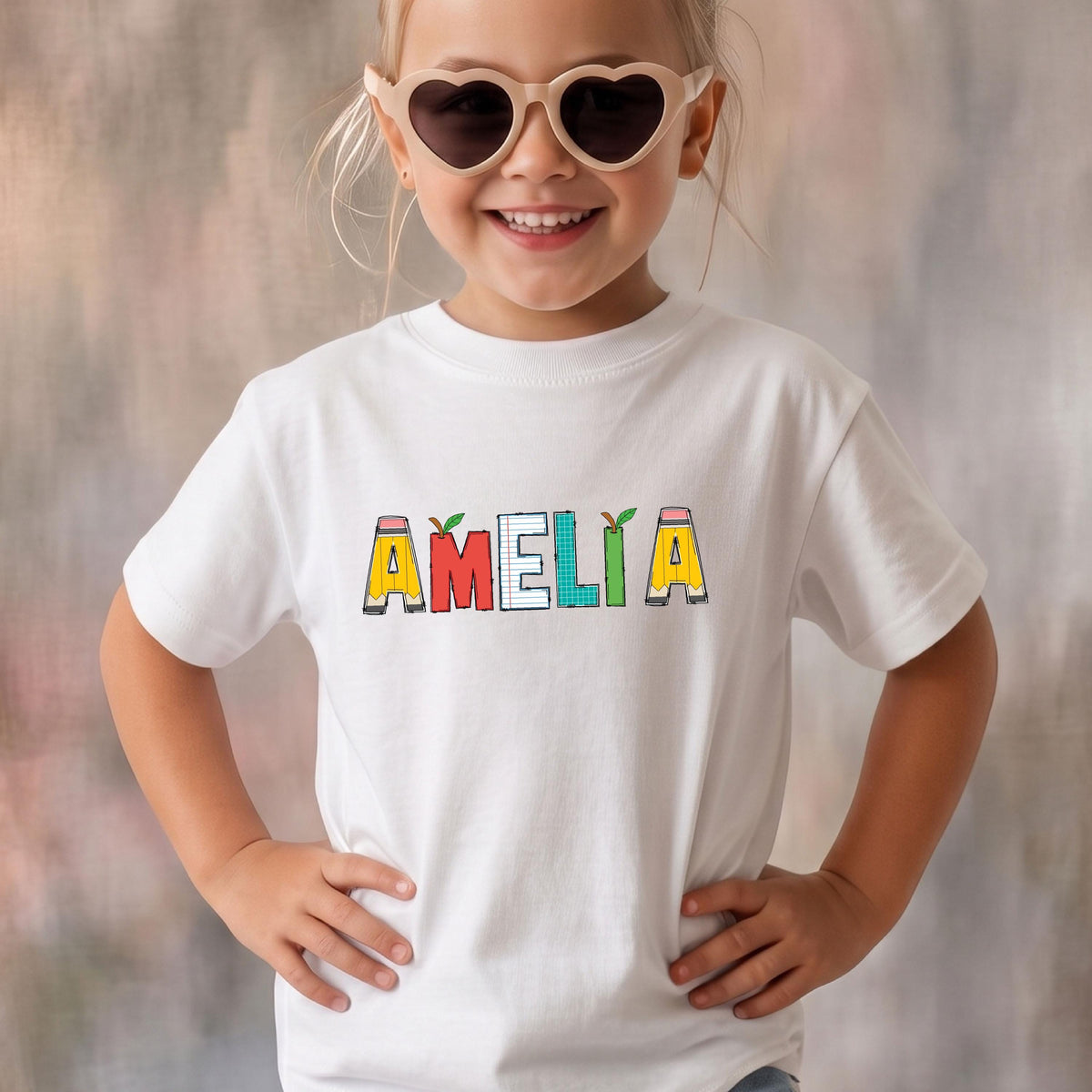 May include: A white short-sleeved t-shirt featuring the name AMELIA printed in a colorful, school-themed font. Each letter is designed to look like a different school supply, including pencils, lined paper, and graph paper patterns.