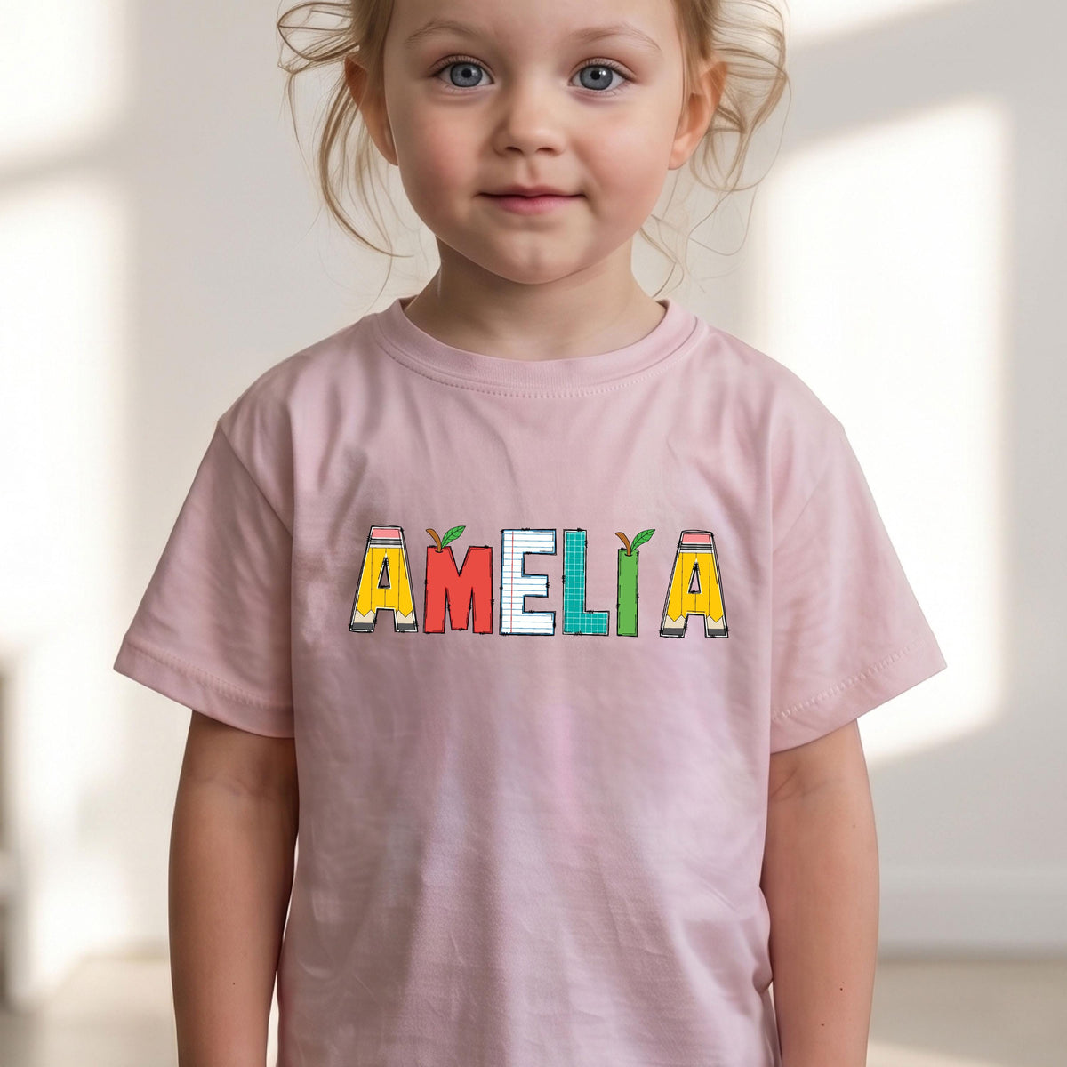 May include: A light pink short-sleeved t-shirt featuring the name AMELIA printed across the chest. The letters are designed with school-themed patterns, including yellow pencils, red apples, lined notebook paper, and green grid paper.