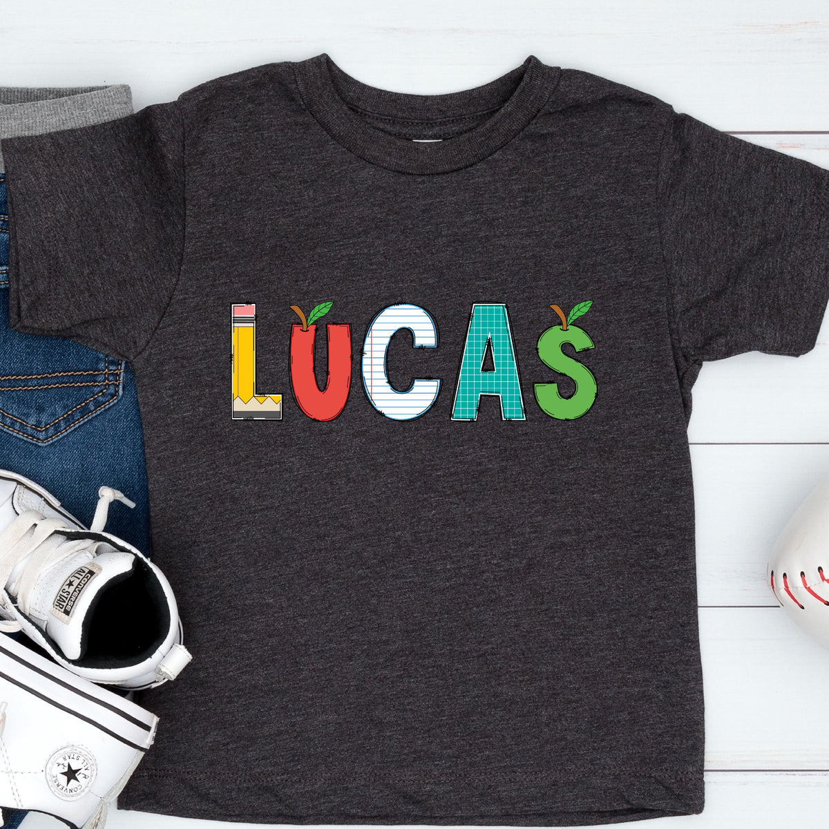 May include: A dark charcoal gray short-sleeve t-shirt featuring the name LUCAS printed in large letters. Each letter has a unique school-themed pattern: L is a yellow pencil, U is a red apple, C is lined paper, A is green graph paper, and S is a green apple.