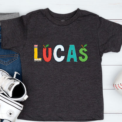 May include: A dark charcoal gray short-sleeve t-shirt featuring the name LUCAS printed in large letters. Each letter has a unique school-themed pattern: L is a yellow pencil, U is a red apple, C is lined paper, A is green graph paper, and S is a green apple.