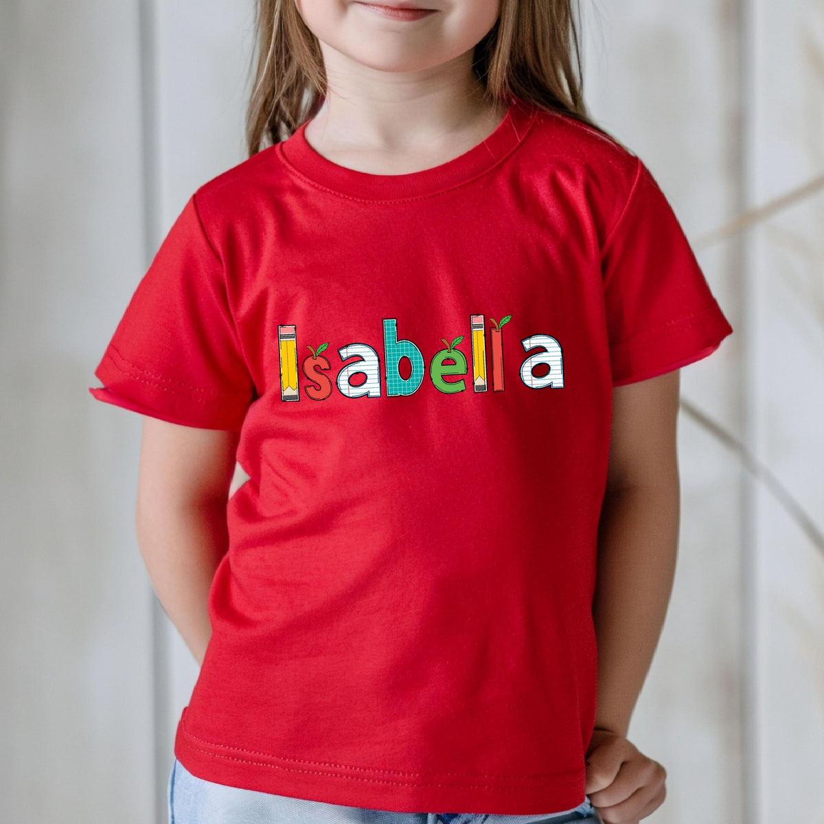 May include: A bright red short-sleeved t-shirt featuring the name Isabella printed across the chest. Each letter is designed with a unique school-themed pattern, including pencils, apples, and grid paper textures in yellow, red, white, green, and orange.