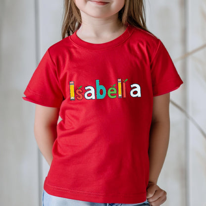 May include: A bright red short-sleeved t-shirt featuring the name Isabella printed across the chest. Each letter is designed with a unique school-themed pattern, including pencils, apples, and grid paper textures in yellow, red, white, green, and orange.
