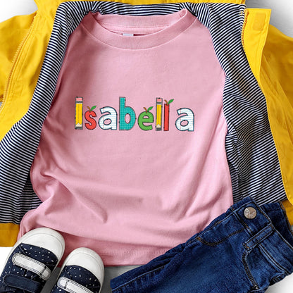 May include: A light pink t-shirt featuring the name Isabella printed in a colorful, school-themed font. The letters are designed with patterns resembling pencils, apples, and lined notebook paper. The shirt is layered under a bright yellow jacket.