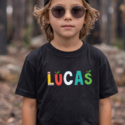 May include: A black short-sleeve t-shirt featuring the name LUCAS printed in colorful, school-themed letters. The L is a yellow pencil, the U is a red apple, the C is white lined paper, the A is a green grid pattern, and the S is a green apple.