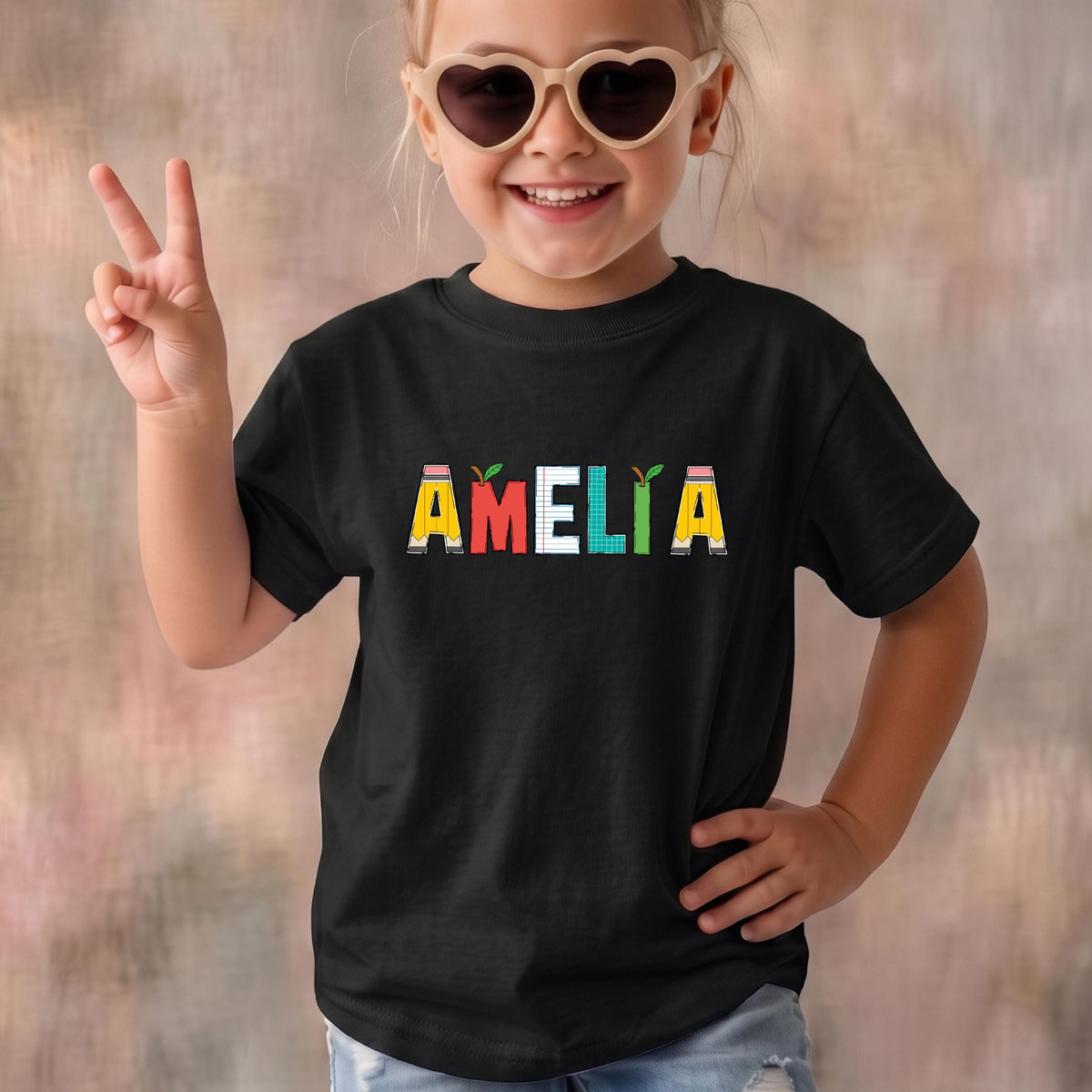 May include: A black short-sleeved t-shirt featuring the name AMELIA printed in large, colorful letters across the chest. Each letter is designed with a unique pattern, including pencil motifs, lined paper textures, and small green apple accents.