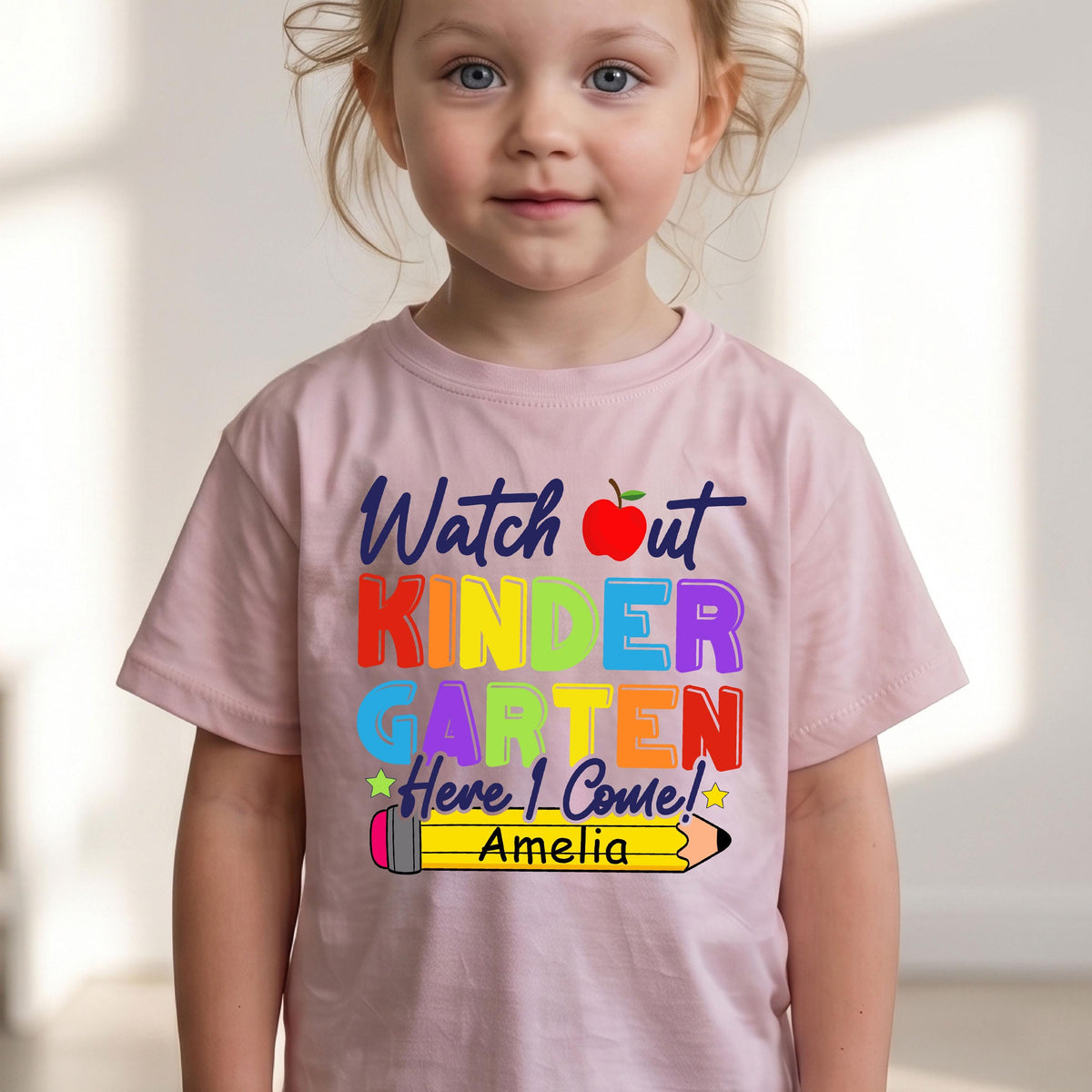 May include: A light pink short-sleeved t-shirt featuring the text "Watch out KINDERGARTEN Here I Come!" in multi-colored letters, a red apple icon, and a yellow pencil graphic with the name "Amelia" printed in black on the pencil body.