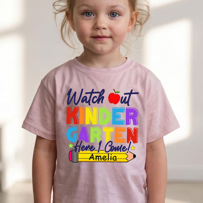 May include: A light pink short-sleeved t-shirt featuring the text "Watch out KINDERGARTEN Here I Come!" in multi-colored letters, a red apple icon, and a yellow pencil graphic with the name "Amelia" printed in black on the pencil body.