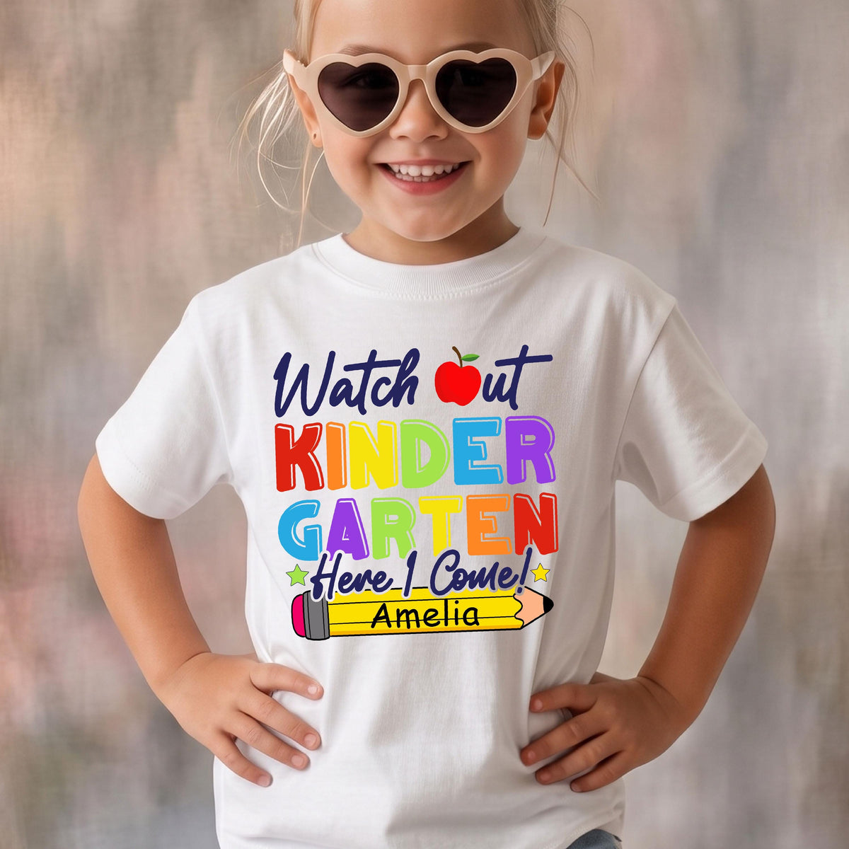 May include: A white short-sleeved t-shirt featuring the text Watch out KINDERGARTEN Here I Come! in colorful letters, a red apple icon, and a yellow pencil graphic with the name Amelia printed in black text on the pencil body.