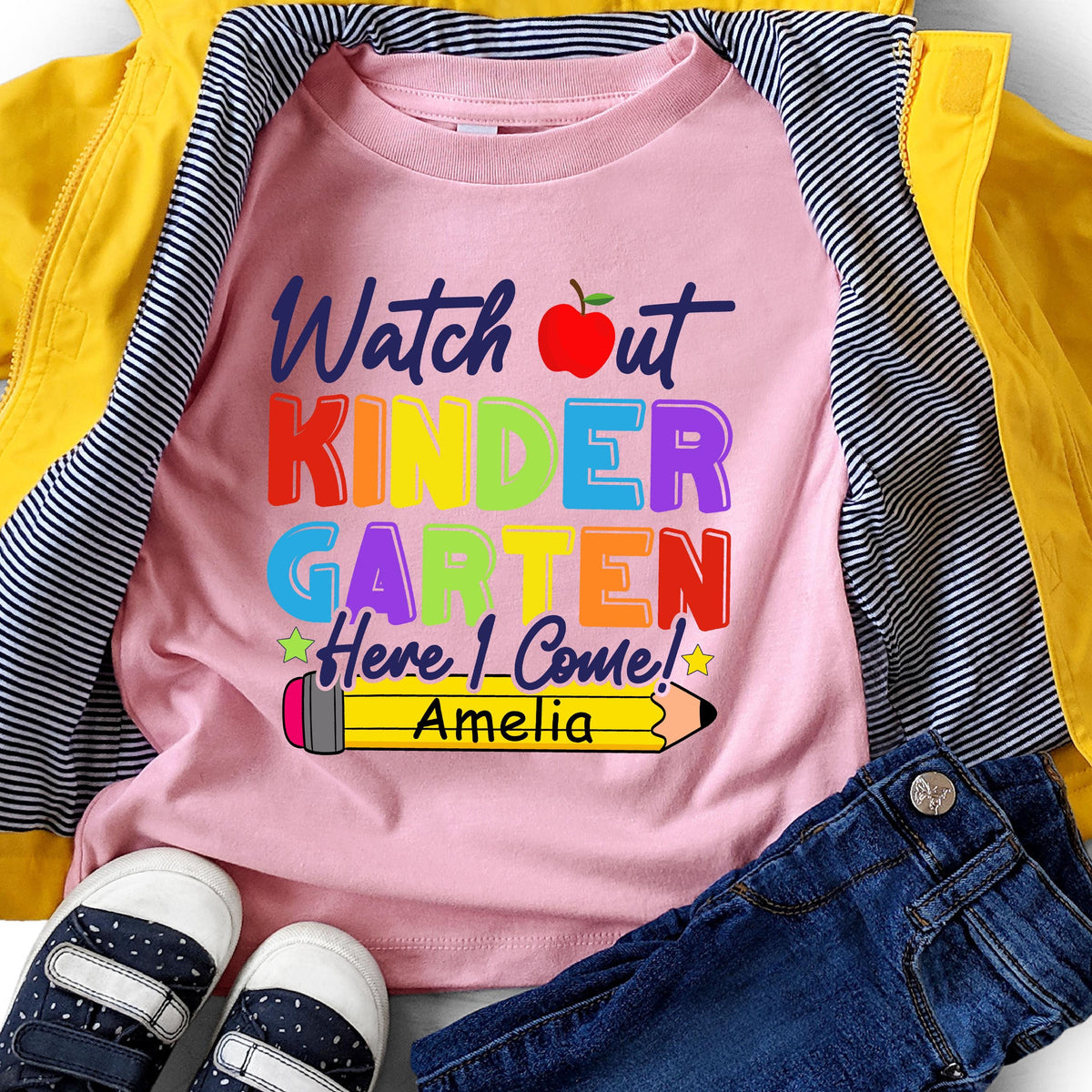 May include: A light pink t-shirt featuring the text Watch out KINDERGARTEN Here I Come! in colorful letters, a red apple graphic, and a yellow pencil illustration with the name Amelia printed in black on the pencil body.