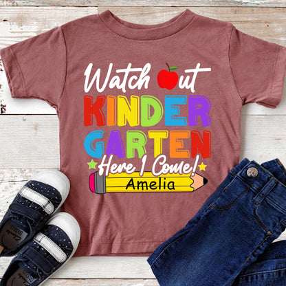 May include: A mauve short-sleeved t-shirt featuring the text Watch out KINDERGARTEN Here I Come! in colorful letters, a red apple graphic, and a yellow pencil graphic with the name Amelia printed in black on the pencil body.