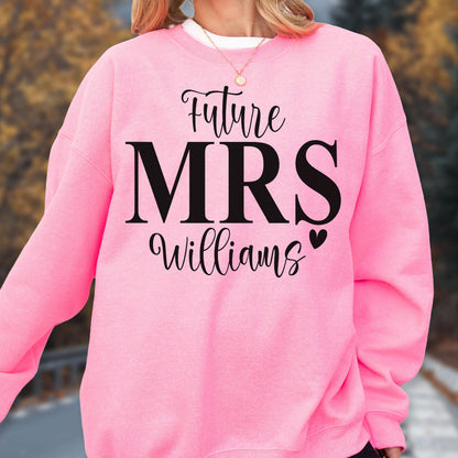 May include: A bright pink crewneck sweatshirt featuring black text that reads Future MRS Williams with a small black heart icon. The garment has long sleeves and a ribbed neckline, showcasing a bold graphic design printed on the front center.