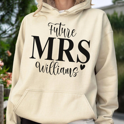May include: A cream-colored hooded sweatshirt featuring black text on the front that reads Future MRS Williams with a small heart icon. The garment includes a front kangaroo pocket, long sleeves, and a drawstring hood with matching cream cords.