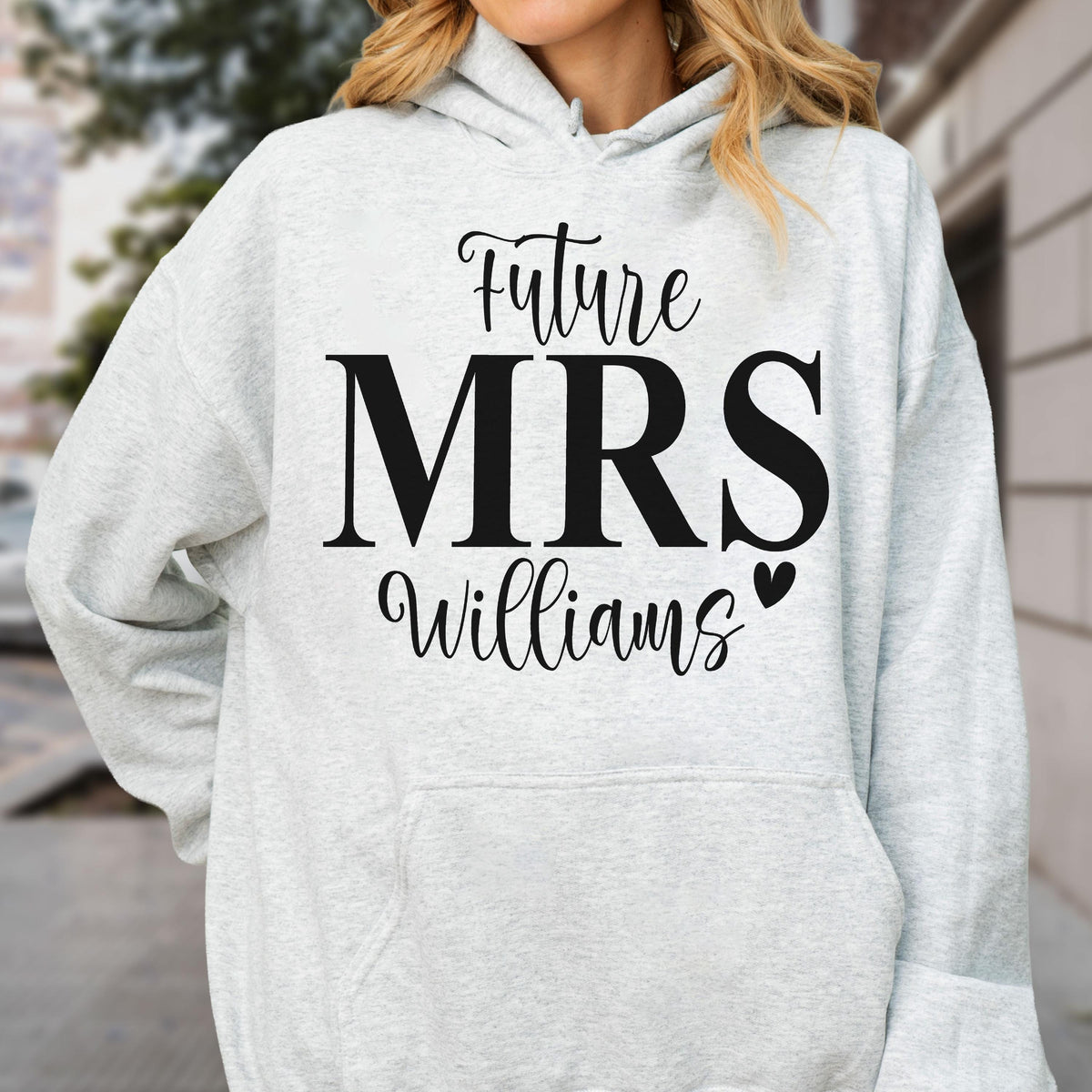 May include: A light gray hooded sweatshirt featuring black text that reads Future MRS Williams with a small black heart icon. The garment includes a drawstring hood and a front kangaroo pocket, showcasing a casual design with long sleeves.