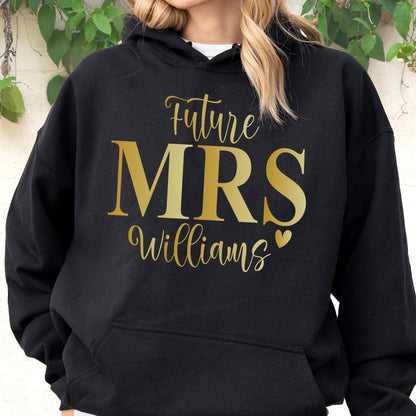 May include: A black hooded sweatshirt featuring gold text that reads Future MRS Williams with a small gold heart icon. The garment includes a front kangaroo pocket, long sleeves, and a drawstring hood, providing a casual and comfortable fit.