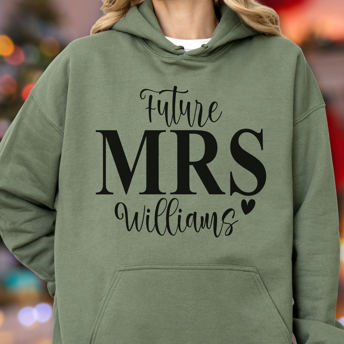 May include: An olive green hooded sweatshirt featuring black text that reads Future MRS Williams with a small heart icon. The garment includes a front kangaroo pocket, long sleeves, and a drawstring hood, providing a casual look with clear lettering.