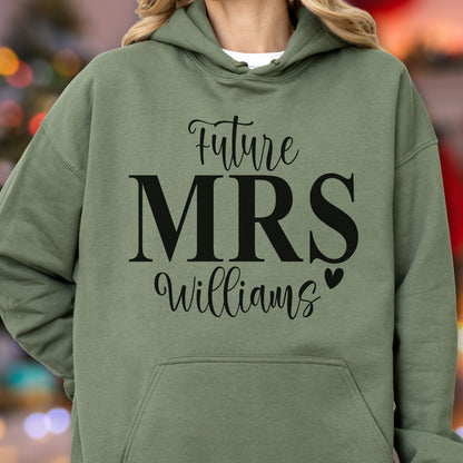 May include: An olive green hooded sweatshirt featuring black text that reads Future MRS Williams with a small heart icon. The garment includes a front kangaroo pocket, long sleeves, and a drawstring hood, providing a casual look with clear lettering.