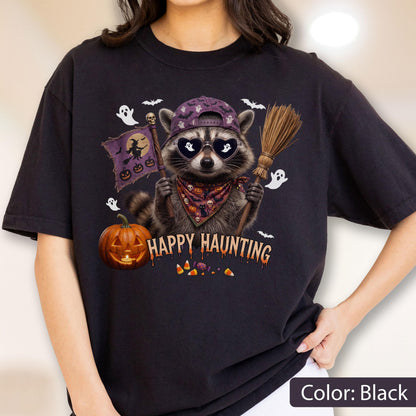 May include: A black t-shirt featuring a graphic of a raccoon wearing a purple baseball cap and heart-shaped sunglasses. The raccoon holds a broom and a flag with a witch silhouette. Text reads HAPPY HAUNTING, surrounded by ghosts, bats, and a pumpkin.