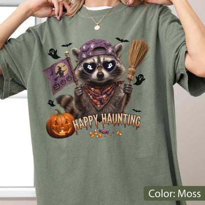 May include: A moss green t-shirt featuring a graphic of a raccoon wearing a purple baseball cap, heart-shaped sunglasses, and a patterned bandana. The raccoon holds a broom and a flag with a witch silhouette. Text reads Happy Haunting.