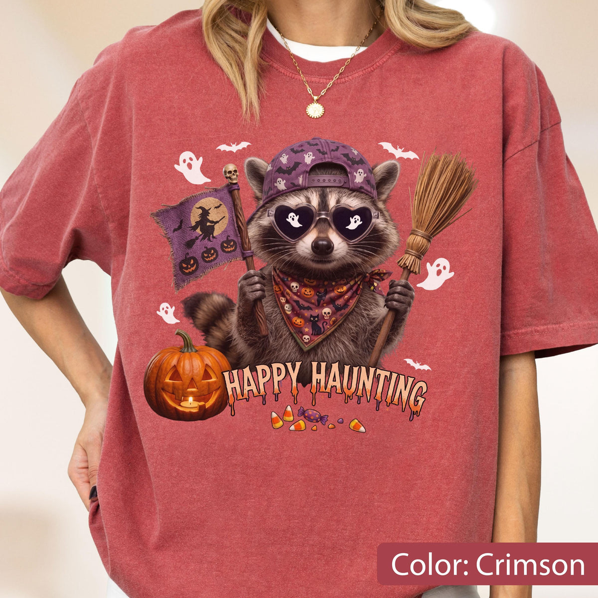 May include: A crimson t-shirt featuring a graphic of a raccoon wearing a purple baseball cap and heart-shaped sunglasses. The raccoon holds a flag with a witch silhouette and a broomstick. Text reads HAPPY HAUNTING above a pumpkin and candy.