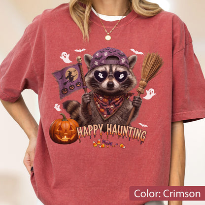 May include: A crimson t-shirt featuring a graphic of a raccoon wearing a purple baseball cap and heart-shaped sunglasses. The raccoon holds a flag with a witch silhouette and a broomstick. Text reads HAPPY HAUNTING above a pumpkin and candy.