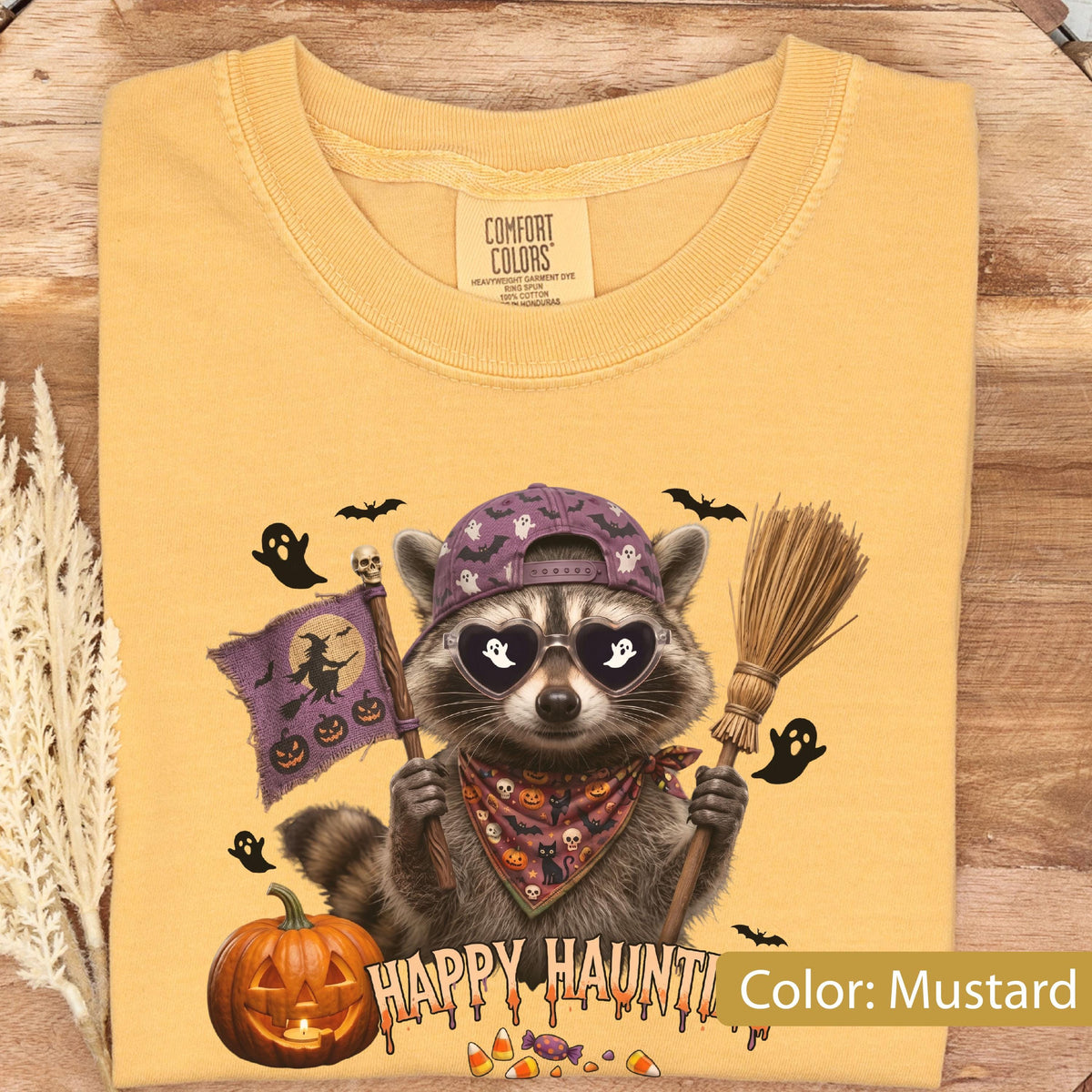 May include: A mustard yellow t-shirt featuring a graphic of a raccoon wearing a purple baseball cap and heart-shaped sunglasses. The raccoon holds a purple flag with a witch silhouette and a broomstick, surrounded by ghosts, bats, and a pumpkin.