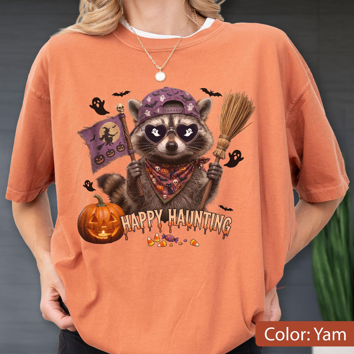 May include: A yam-colored t-shirt featuring a graphic of a raccoon wearing a purple baseball cap and heart-shaped sunglasses. The raccoon holds a broom and a purple flag with a witch silhouette. Text reads Happy Haunting with pumpkins and candy.