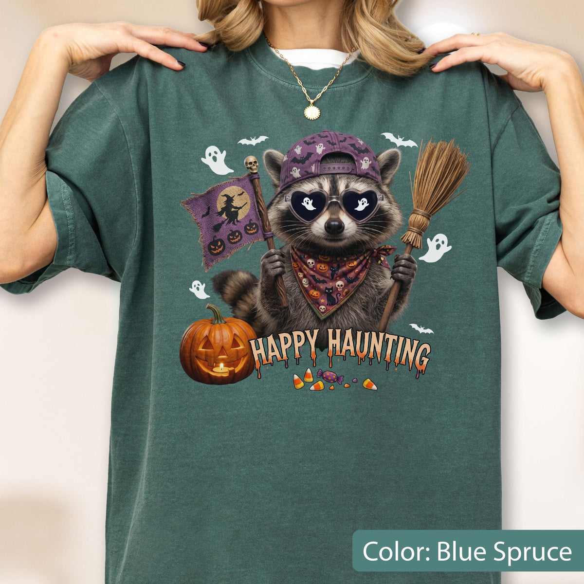 May include: A blue spruce t-shirt featuring a graphic of a raccoon wearing a purple cap, heart-shaped sunglasses, and a patterned bandana. The raccoon holds a broom and a flag with a witch silhouette. Text reads HAPPY HAUNTING with pumpkins.